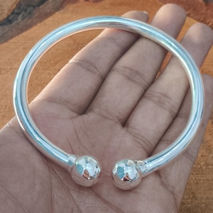 Puede incluir: Una pulsera abierta de tono plateado pulido que presenta una banda circular lisa que termina en ambos extremos con un adorno esférico. La superficie metálica es reflectante y de color uniforme, creando una forma circular simple y continua.