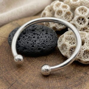 May include: A polished silver cuff bracelet with a rounded design and two ball ends. The bracelet is displayed on a wooden surface with a black stone and coral in the background. The bracelet is a simple, elegant piece of jewelry.
