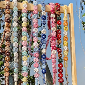 Può includere: Una collezione di otto lunghe cinghie verticali realizzate con motivi floreali all'uncinetto, ciascuna dotata di una perlina bianca centrale. Le cinghie presentano varie combinazioni di colori tra cui marrone, azzurro, giallo, rosa, blu scuro, rosso e arancione.