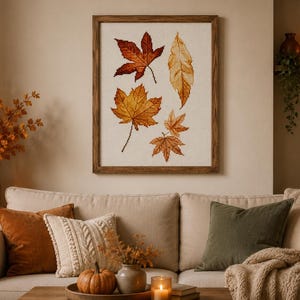 May include: A framed cross-stitch embroidery featuring four distinct autumn leaves in shades of deep red, orange, and golden yellow on a cream-coloured fabric background, enclosed within a simple dark brown wooden frame hanging on a wall.