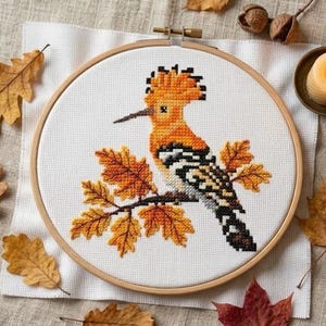 May include: A circular wooden embroidery hoop containing a cross-stitch pattern of a hoopoe bird with orange, black, and white plumage perched on a branch with orange autumn leaves, set against a white fabric background with a visible grid.