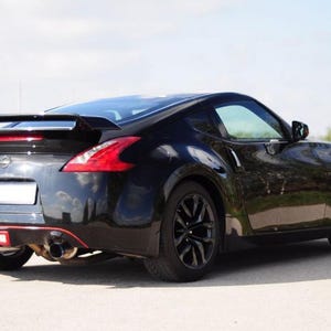 Pode incluir: Um cupê esportivo Nissan 370Z preto visto de um ângulo traseiro de três quartos, apresentando um aerofólio traseiro, rodas de liga leve escuras, escapamento duplo e uma linha vermelha de destaque ao longo do para-choque inferior, estacionado em uma superfície pavimentada.