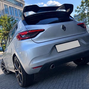 May include: A silver Renault hatchback viewed from the rear, featuring a prominent black roof spoiler, dark multi-spoke alloy wheels, red-tinted taillights, a central diamond-shaped emblem, and a single exhaust pipe integrated into the black bumper.