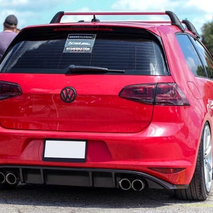Könnte beinhalten: Ein leuchtend roter Volkswagen Schrägheck von hinten gesehen, mit schwarzem Emblem, getönten Rückleuchten, Dachträger und Heckdiffusor mit vier Auspuffendrohren. Die Heckscheibe zeigt drei Aufkleber, darunter einen mit der Aufschrift Stancenation.