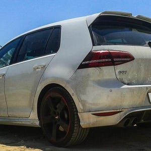 Könnte beinhalten: Ein weißer Volkswagen Golf Schrägheck aus der hinteren Dreiviertelansicht. Das Fahrzeug verfügt über einen Heckspoiler, dunkle Fünfspeichen-Leichtmetallfelgen mit roten Akzenten, einen schwarzen Heckdiffusor, zwei Auspuffrohre und ein Volkswagen-Logo.