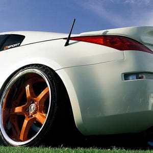 Könnte beinhalten: Ein weißer Sportwagen mit Heckspoiler, schwarzer Antenne und leuchtend orangefarbenen Fünfspeichenfelgen mit polierten Rändern. Das Fahrzeug hat ein tiefes Profil, ein dunkel getöntes Fenster mit K-SPORT-Aufkleber und ein großes Auspuffrohr.