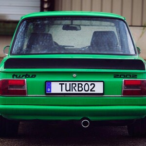 Peut inclure: Une voiture ancienne vert vif vue de l'arrière, dotée d'un aileron noir, de feux arrière rectangulaires rouges et blancs, et d'une plaque d'immatriculation indiquant TURB02. La voiture possède un pot d'échappement central et l'inscription turbo 2002.