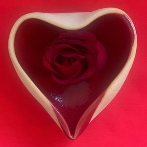 May include: A heart-shaped ceramic bowl with a glossy deep red interior and a matte beige exterior rim. A single dark red rose blossom is placed inside the bowl, resting against the curved bottom surface of the vessel.