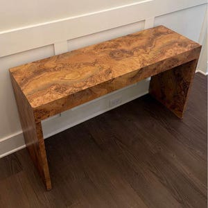 Könnte beinhalten: Eine rechteckige Holzbank mit einem einzigartigen Maserungsmuster. Die Bank hat eine warme, natürliche Farbpalette mit Braun- und Beigetönen. Sie hat ein schlichtes, modernes Design mit einer flachen Oberseite und zwei Seitenstützen.