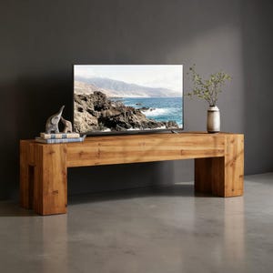 May include: A rectangular light brown wooden media console with a thick, block-style frame. On top sits a flat-screen television displaying a coastal landscape, two small gray elephant figurines on stacked books, and a white vase with green foliage.