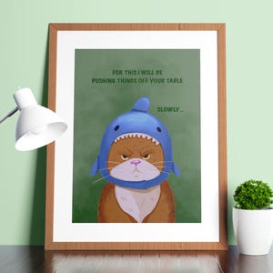 May include: A framed wall art print featuring an illustration of an orange cat wearing a blue shark costume. The text reads: FOR THIS I WILL BE PUSHING THINGS OFF YOUR TABLE SLOWLY. The cat has a grumpy expression, and the background is a solid green.