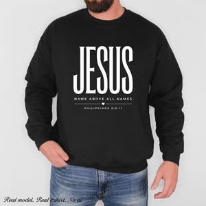 以下が含まれることがあります： 胸元に白い文字で「JESUS NAME ABOVE ALL NAMES」とプリントされ、小さなハートのアイコンと「PHILIPPIANS 2:9-11」という聖書の引用が記された黒いクルーネックのスウェットシャツ。長袖で、袖口と裾はリブ編みになっています。