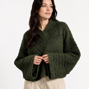 May include: A dark green, open-front sweater with a textured knit pattern. The sweater features wide, bell sleeves and a V-neck design. The model is wearing the sweater with beige shorts.