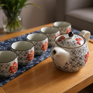 Peut inclure: Service à thé en céramique composé d'une théière et de tasses assorties. La théière et les tasses sont blanches avec un motif floral rouge et noir. L'ensemble est disposé sur une surface en bois.