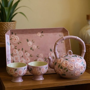 Könnte beinhalten: Ein Teeservice bestehend aus einer Teekanne mit bambusartigem Henkel, zwei Teetassen mit Fuß und einem rechteckigen Serviertablett. Jedes Teil besteht aus rosa Keramik mit einem handgemalten Muster aus hellrosa Rosen und grünen Blättern.