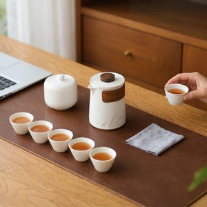 Può includere: Un set da tè in ceramica bianca composto da una teiera con coperchio e fascia in legno, un contenitore con coperchio e cinque piccole tazze da tè riempite di liquido ambrato. La base della teiera presenta un rilievo di paesaggio montano.
