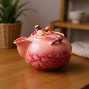 May include: A pink ceramic teapot with a floral design. The teapot has a curved spout, a rounded body, and a lid with decorative accents. The floral pattern is a darker shade of pink, and the accents are gold and purple.