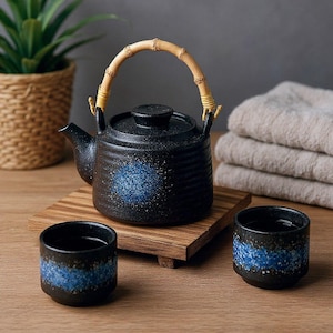 May include: A black ceramic teapot with a curved bamboo handle and a matching lid, featuring a textured blue speckled circular design. Two small black ceramic cups with a horizontal blue speckled band accompany the teapot on a wooden base.