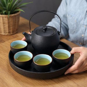 Puede incluir: Una tetera de hierro fundido negro con un asa curva y una tapa, acompañada de cuatro tazas de té pequeñas sobre una bandeja negra. Las tazas tienen un interior azul claro y un borde dorado, llenas de té verde.