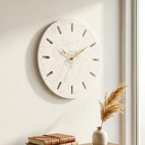 May include: A circular wall clock with a textured off-white surface and gold-colored hands. The clock face features elongated rectangular cutouts as hour markers instead of numbers, creating a clean and simple design for any interior wall space.