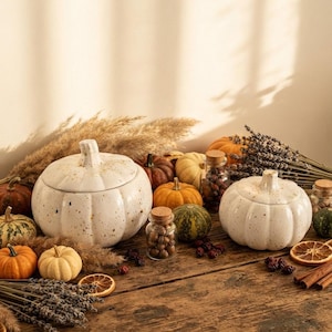 May include: Two white ceramic pumpkin-shaped lidded containers with brown speckles, surrounded by small real pumpkins, dried lavender bundles, pampas grass, glass jars filled with dried berries and nuts, and dried orange slices on a wooden surface.