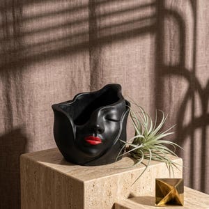 Peut inclure: Un pot de fleurs en céramique noire en forme de visage aux yeux fermés et aux lèvres rouges. Le pot est posé sur un bloc de pierre clair, avec une petite sculpture géométrique dorée et une plante verte à proximité. Le fond est un tissu texturé de ton neutre.