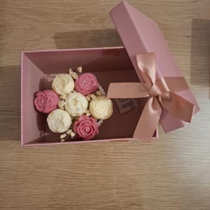 Peut inclure: Une boîte cadeau rose avec un nœud en ruban de satin assorti. À l'intérieur, plusieurs fleurs de savon roses et blanches sont disposées avec de petites fleurs de remplissage blanches. La boîte est ouverte.