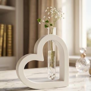 Op de afbeelding: Een witte hartvormige vaas houdt een heldere glazen buis met water en kleine witte bloemen vast. De vaas is gemaakt van een effen, mat materiaal en staat op een marmeren oppervlak. De achtergrond omvat een raam en een vage plank met boeken.
