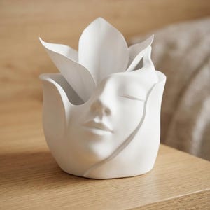 Peut inclure: Un pot de fleurs en céramique blanc mat en forme de visage humain émergeant d'une fleur. La sculpture présente des yeux fermés, un nez et des lèvres intégrés dans les pétales incurvés qui forment le récipient, créant un objet sculptural lisse.