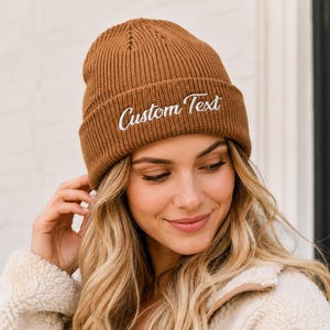 May include: A ribbed knit beanie in a solid brown colour featuring the words Custom Text embroidered in white cursive script across the front cuff. The hat has a classic folded brim design and a textured vertical knit pattern throughout the fabric.