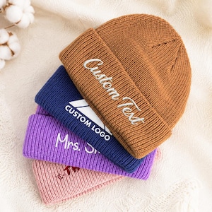 May include: A stack of four cuffed knit beanies in brown, navy blue, purple, and light pink. The brown beanie features white embroidered text reading Custom Text. The navy blue beanie has a white logo reading CUSTOM LOGO. The purple beanie has white text reading Mrs. S. The pink beanie has a small dark graphic.