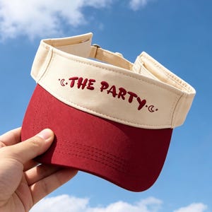 Op de afbeelding: Een tweekleurige zonneklep met een crèmekleurige voorband voorzien van de rode geborduurde tekst THE PARTY en kleine halve maan- en sterpictogrammen, gecombineerd met een donkerrode klep en een verstelbare metalen gespsluiting.