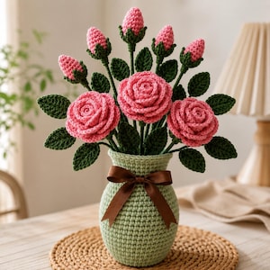 May include: A crochet bouquet featuring three fully bloomed pink roses and six pink rosebuds with dark green leaves, arranged in a light green crocheted vase. The vase is decorated with a brown satin ribbon tied in a bow around the neck.
