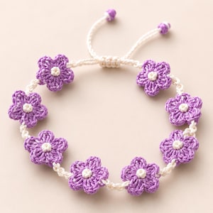 May include: A handmade bracelet featuring crocheted purple flowers with white centers. The flowers are connected by a white cord, and the bracelet has an adjustable closure. The bracelet is a delicate piece of jewelry.