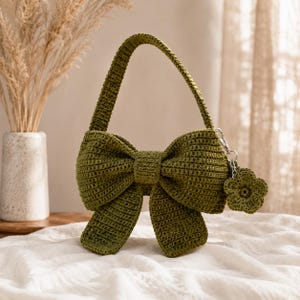 May include: An olive green crochet handbag shaped like a large bow with a single handle. A small, matching crochet flower charm is attached to the side with a silver-toned metal lobster clasp, adding a decorative detail to the textured yarn construction.