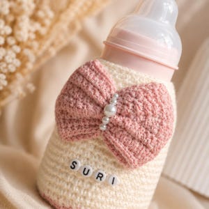 May include: A baby bottle with a cream-colored crocheted cover and a pink bow. The bow is adorned with pearl beads. The word "SURI" is spelled out in small letter beads on the cover.