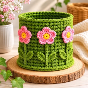 Puede incluir: Una cesta verde de ganchillo con tres aplicaciones de flores rosas y amarillas. La cesta hecha a mano se asienta sobre una rodaja de madera, mostrando un diseño floral encantador. La cesta es un artículo de almacenamiento decorativo.