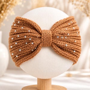 May include: A brown knitted headband with a bow design, adorned with small, sparkling rhinestones. The headband is displayed on a white mannequin head, showcasing its texture and style. This accessory is perfect for adding a touch of elegance to any outfit.