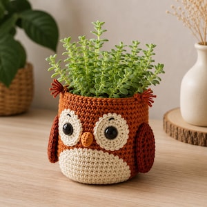 Peut inclure: Un pot de plante en forme de hibou crocheté, aux couleurs marron, crème et orange, rempli de plantes vertes. Le hibou présente de grands yeux ronds, un petit bec et des touffes d'oreilles. Ce pot mesure environ 15 cm de haut.