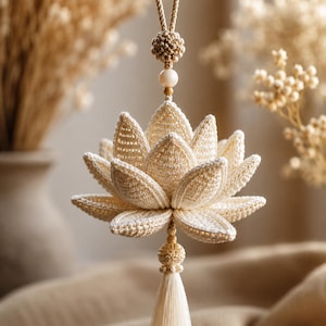 May include: A cream-coloured, crocheted lotus flower ornament with a tassel. The handmade decoration features detailed petal work and a knotted cord for hanging. The ornament is a delicate, handcrafted piece.