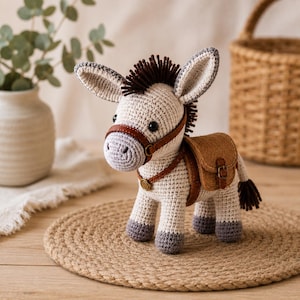 May include: A crocheted donkey figurine with a white body, gray hooves, and a brown mane and tail. It has a brown leather harness and saddlebags. The donkey is standing on a round, woven mat.