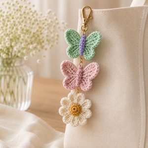 May include: A crochet keychain featuring a green and purple butterfly, a pink butterfly, and a white daisy with a yellow center. The charm is attached to a gold-colored clasp, perfect for a bag or keys.