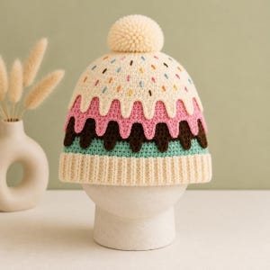 May include: A cream-colored knit hat with a whimsical ice cream cone design. The hat features layers of pink, brown, and teal, with colorful sprinkles and a cream-colored pom-pom on top. The hat is displayed on a white stand.