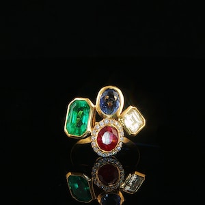 Puede incluir: Un anillo de oro con un conjunto de cuatro piedras preciosas distintas: una esmeralda rectangular, un zafiro azul ovalado, un diamante cuadrado y un rubí ovalado central rodeado por un halo de pequeños diamantes blancos en una banda de oro.