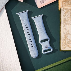Puede incluir: Una correa de reloj de silicona azul claro con motivos celestiales y florales grabados, que incluyen una luna creciente, estrellas y rosas. La correa incluye el texto: "To the stars who listen and the dreams that are answered."