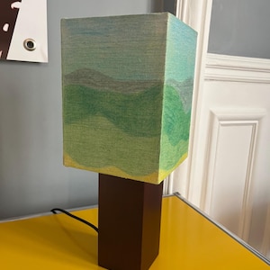 May include: A rectangular table lamp with a brown wooden base and a square lampshade. The lampshade features a landscape painting in shades of green, blue, and yellow. The lamp is plugged into an electrical outlet.