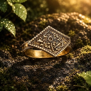 May include: A gold-toned metal ring featuring a prominent diamond-shaped centrepiece. The surface of the diamond is decorated with an intricate, raised relief pattern of swirling lines and small circular motifs, framed by a textured border.