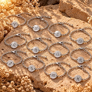 May include: A collection of twelve silver-toned metal chain bracelets, each featuring a circular charm engraved with a unique zodiac sign symbol. The bracelets are arranged in a group, showcasing the twelve astrological signs from Aries to Pisces.