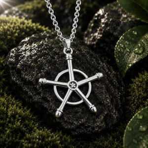 Puede incluir: Un colgante de metal plateado con un marco circular atravesado por tres barras rectas que se extienden hacia afuera, creando una forma de estrella. Una pequeña estrella de cinco puntas está centrada donde las barras tocan el círculo.