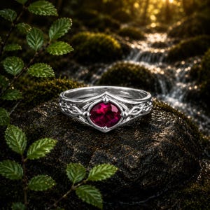 Könnte beinhalten: Ein silberner Ring mit einem zentralen, oval geschliffenen, tiefroten Edelstein. Das Metallband weist an den Seiten komplizierte, symmetrische Filigrandetails auf, die den Stein in einer spitzen, kronenartigen Fassung auf einer dunklen Oberfläche halten.